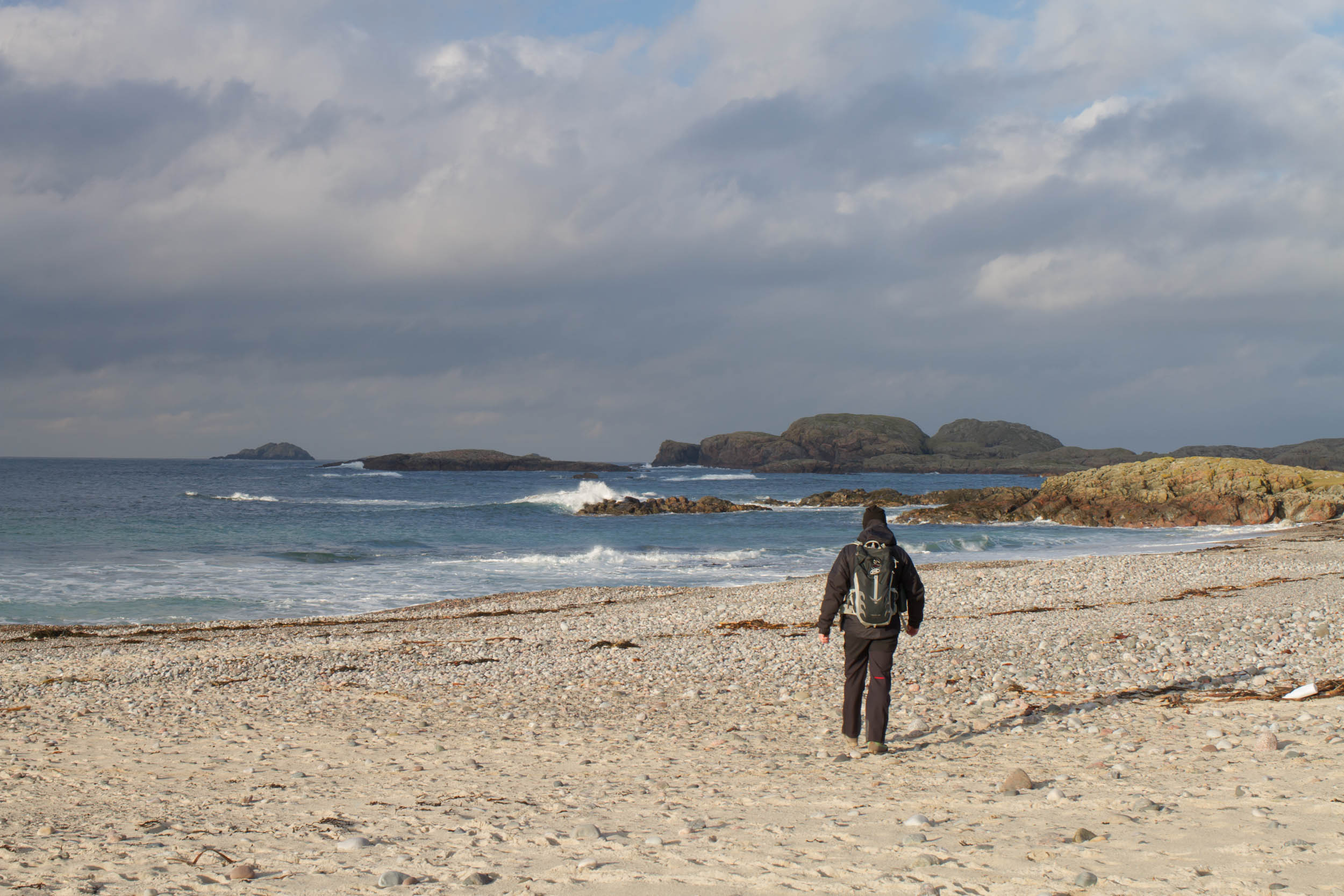 Photo of a man walking across the beach. The sun is shining and the waves are crashing on the rocks