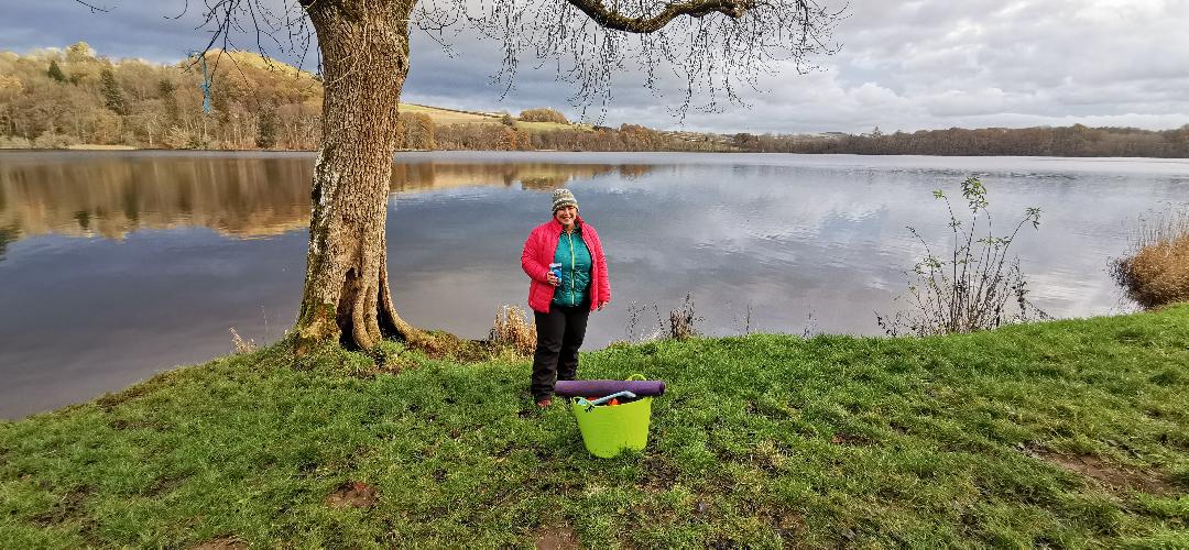 Photo of a woman stood at the side of a loch. Wearing lots of layers after swimming in cold water