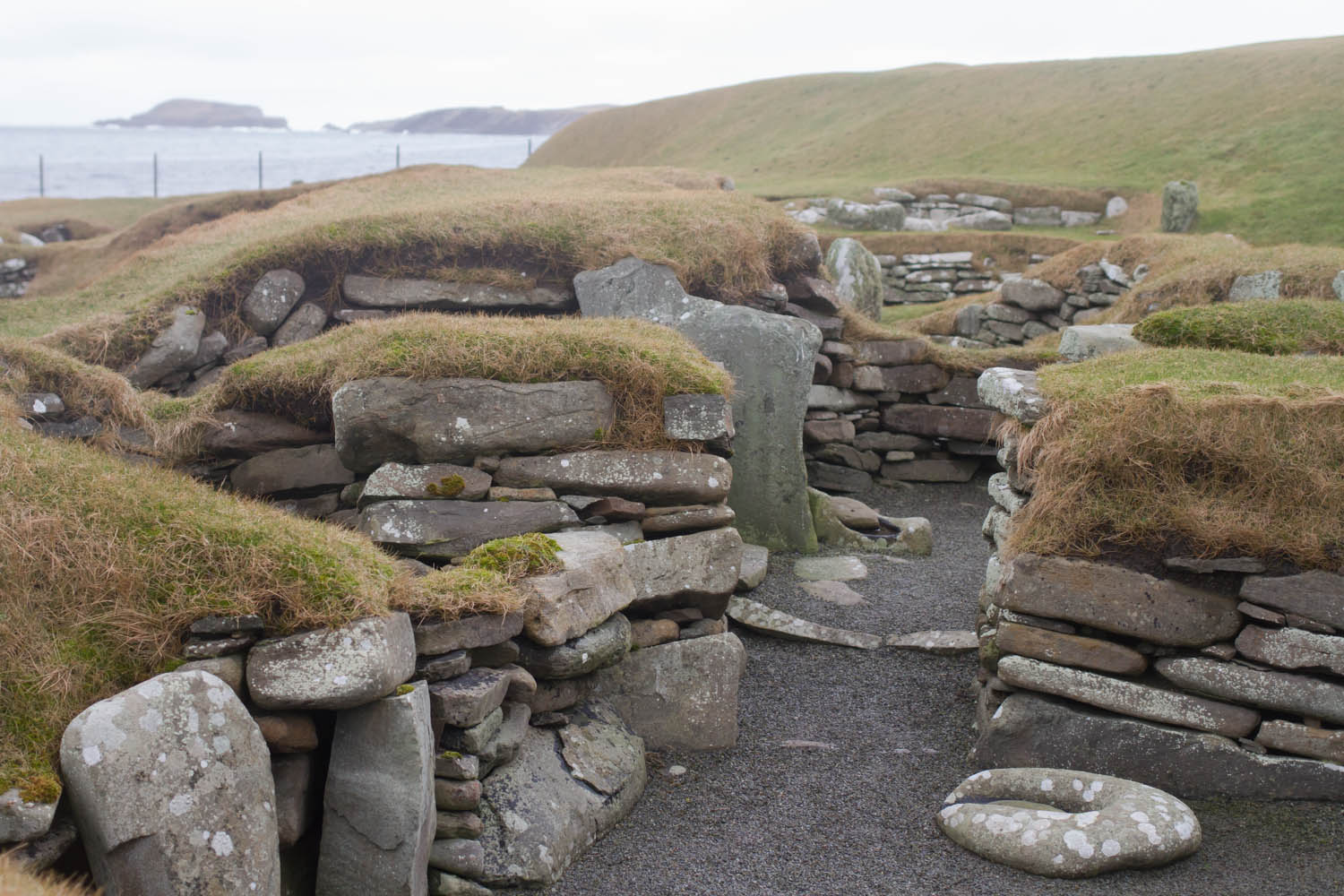 A photo of Jarlshof, Shetland. Showing the layers of stones with grass on top. Pathways which would have formed part of the house  