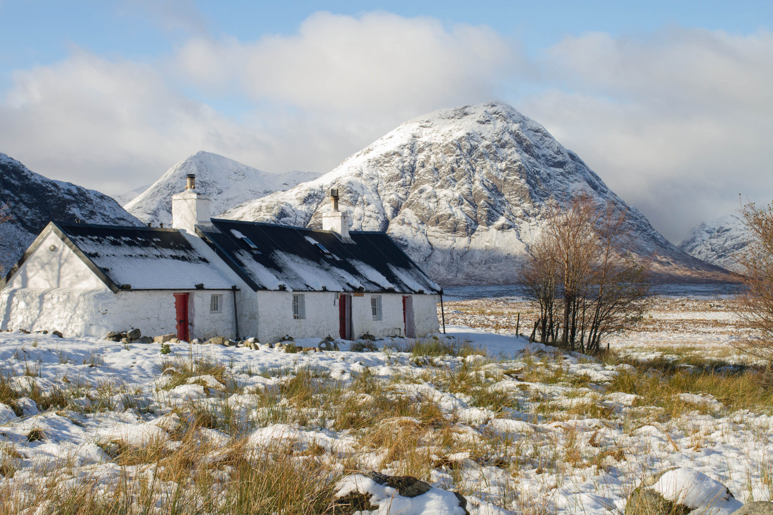 Photo of Buachaille Etive Mor. Snow capped pointy mountain in the background. A small white cottage in the foreground with snow in the foreground