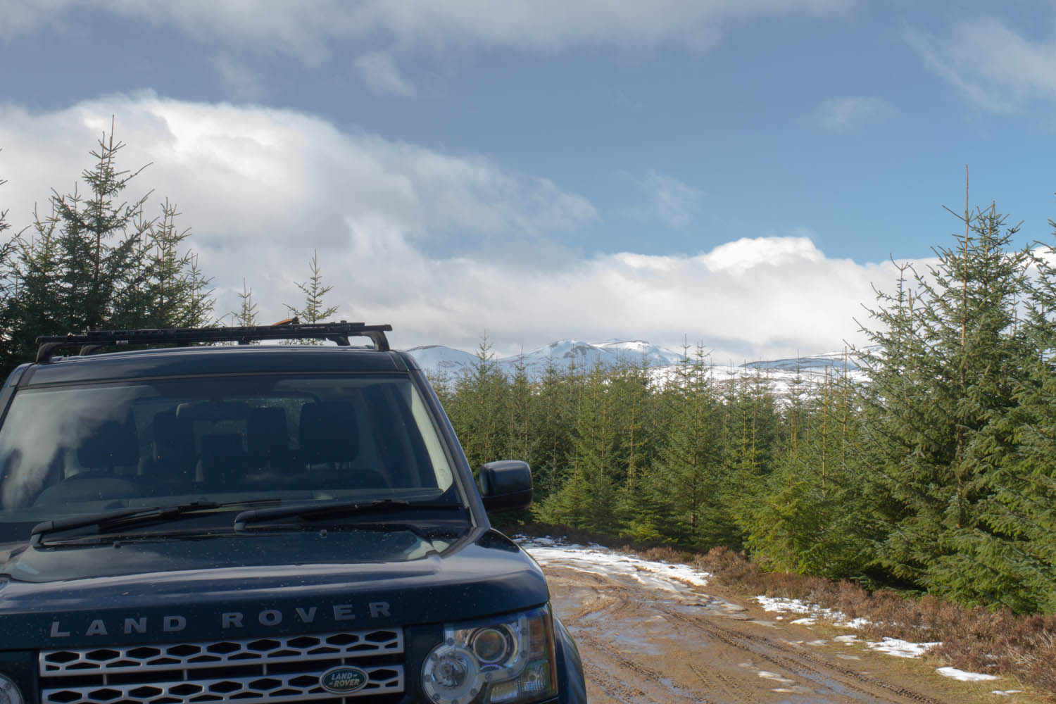 Highland Safaris, Perthshire. Land Rover parked up beside a muddy track. Trees on the side and mountains in the background. Blue sky and big clouds