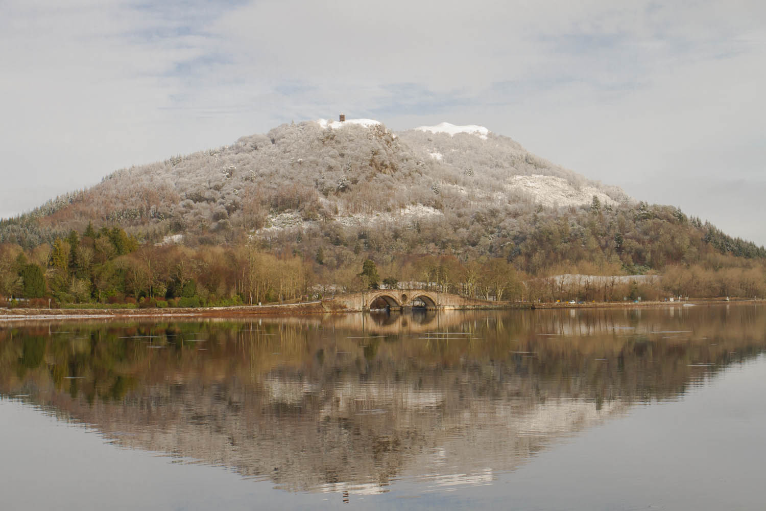 Inveraray, Argyll, Loch Fyne, Scotland. A dusting of snow on the hill. The trees are bare as it is winter. There is a small bridge over the water and the reflection in the foreground is perfectly still