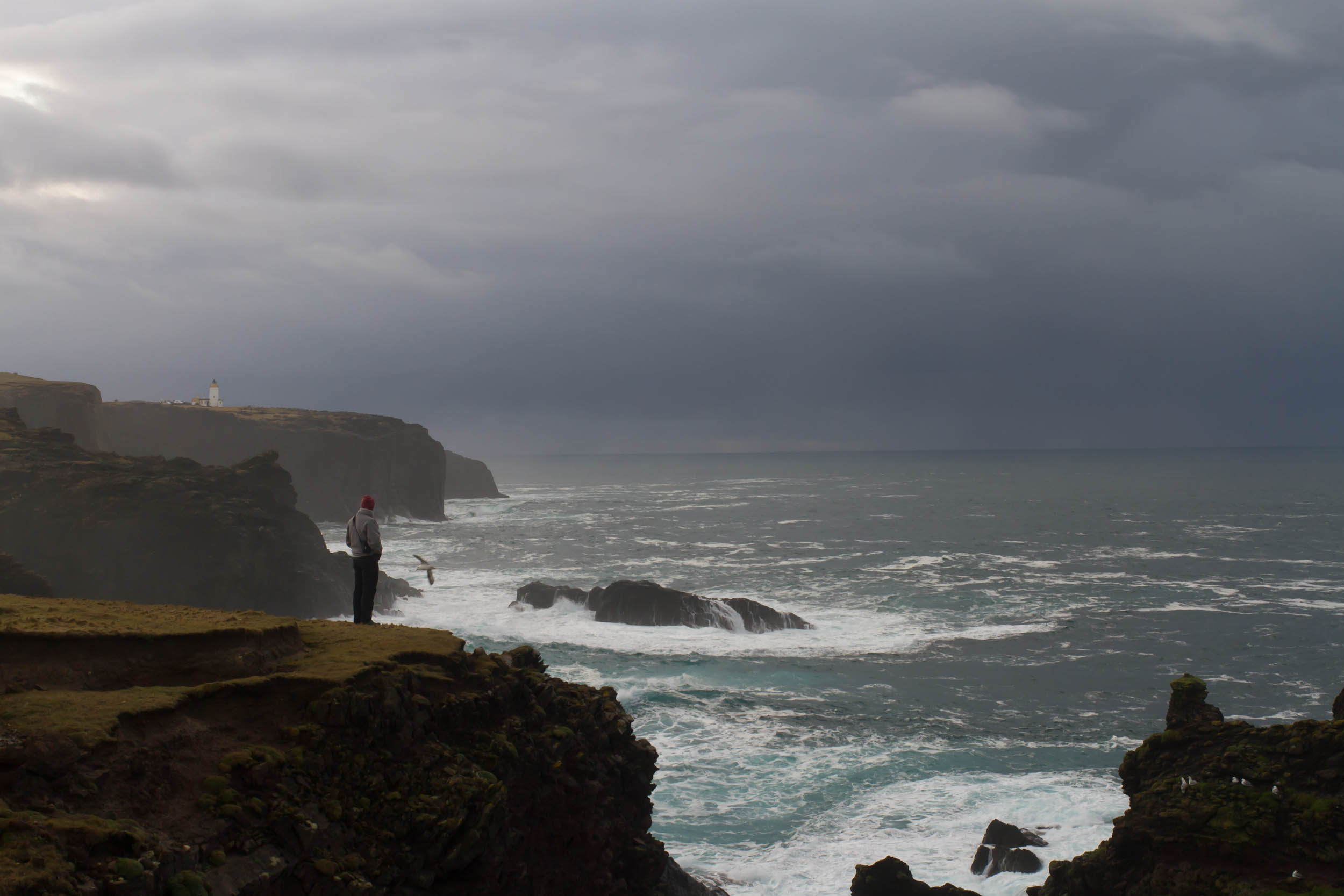 Esha Ness cliffs, Shetland. Man stood on the cliffs. Moody sky, lighthouse in the distance. The sea is crashing against the rocks