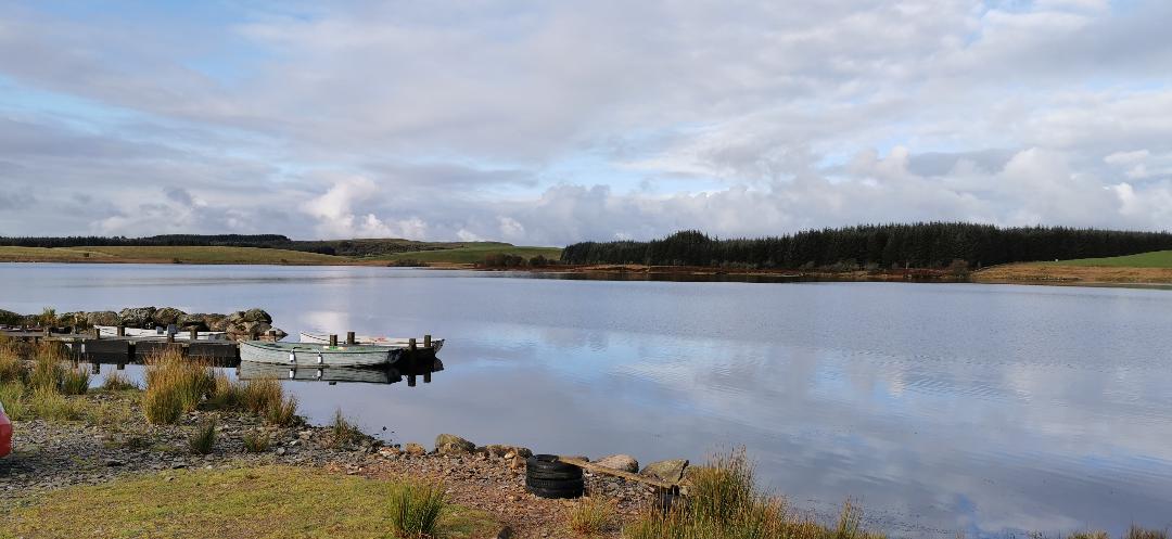 Image of a loch. Clouds reflecting in the sky and a small jetty with boats moored. Wild Swimming at Creeside Escape 